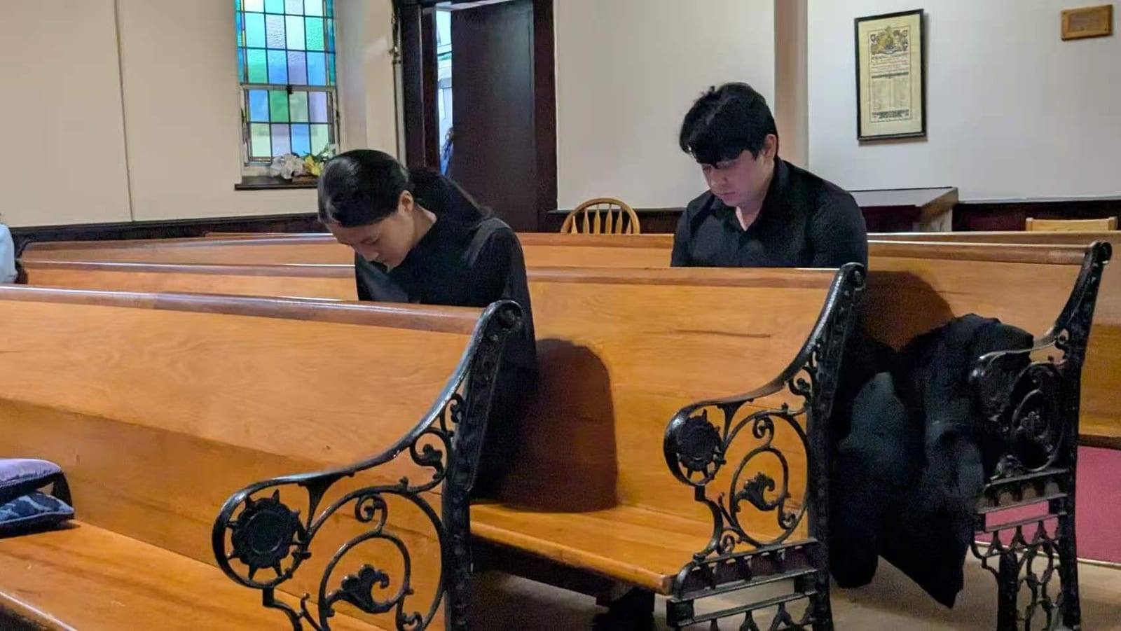 Two students in concert dress sitting quietly in wooden pews, heads down, before going on to compete
