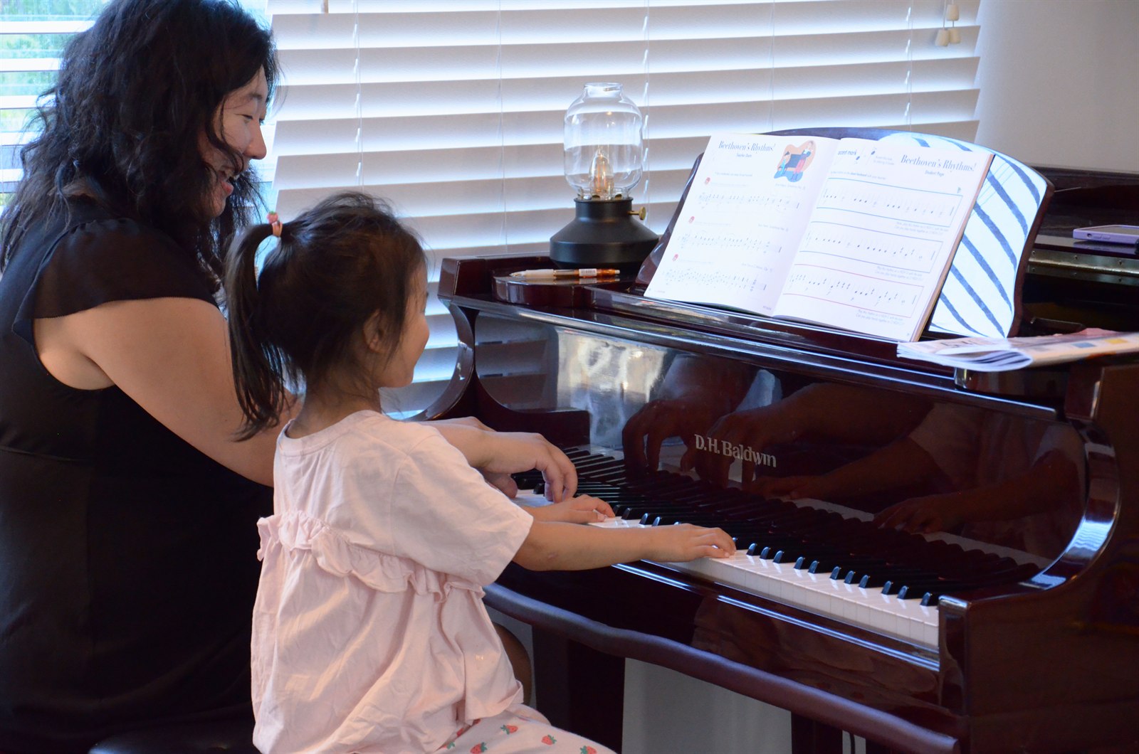 A young beginner reaching for the keys of the grand piano with both hands while her teacher sits beside her, a rhythm workbook open on the music stand