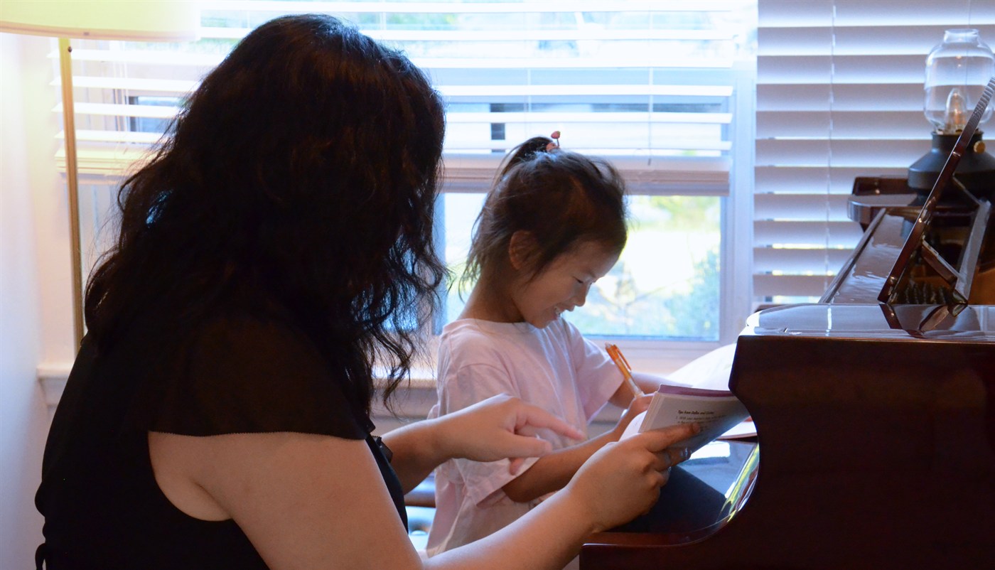 A young student smiling as she works through a lesson booklet at the grand piano, pen in hand, her teacher holding the page open beside her