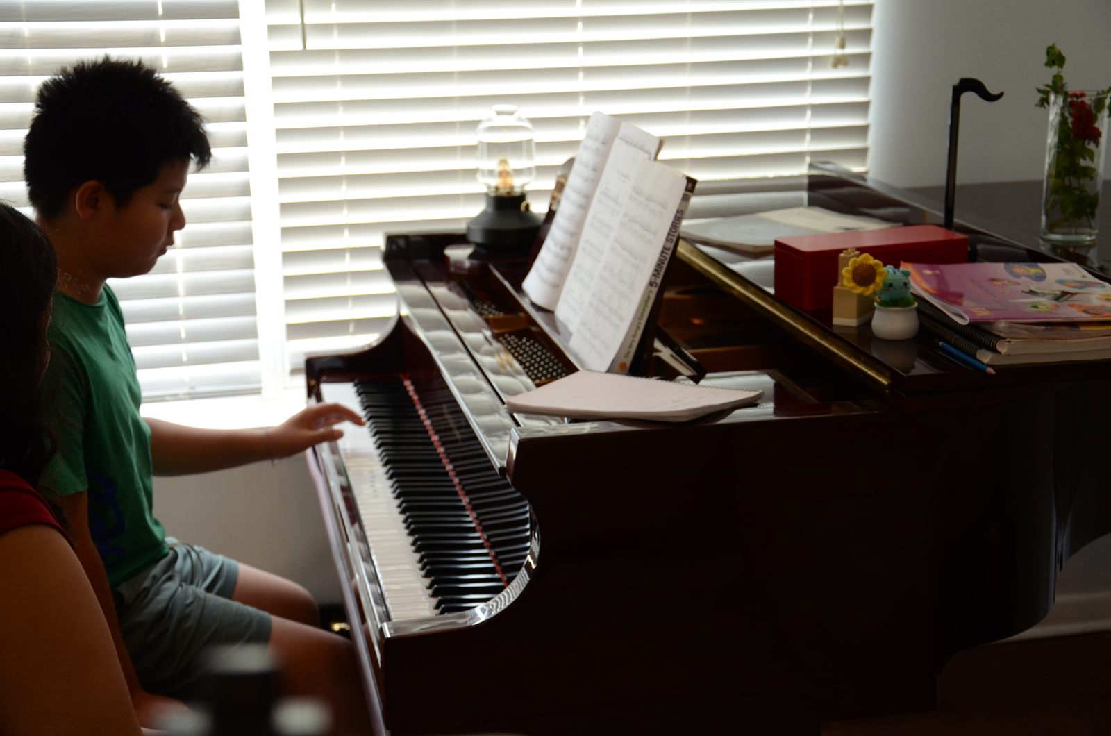 A student in a green shirt playing the grand piano during a lesson, sheet music open on the stand and afternoon light coming through the blinds