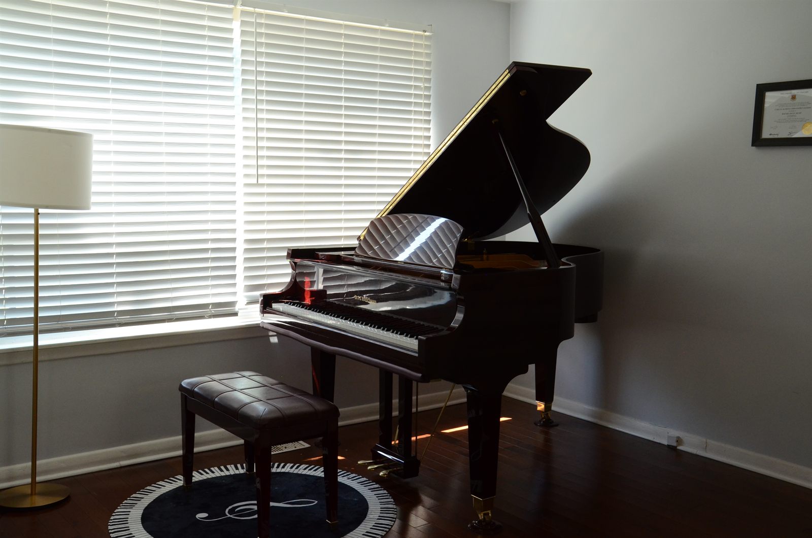 The studio grand piano beside the window, lid raised, with the bench and piano rug in front of it